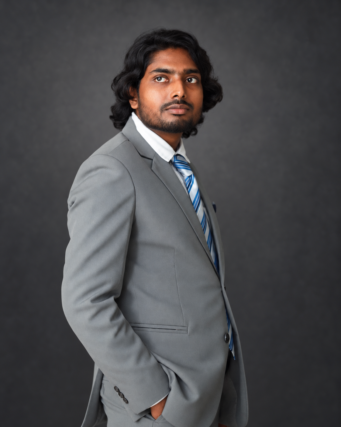 Abdullah Al Tashdid in a grey suit and blue striped tie against a charcoal studio backdrop